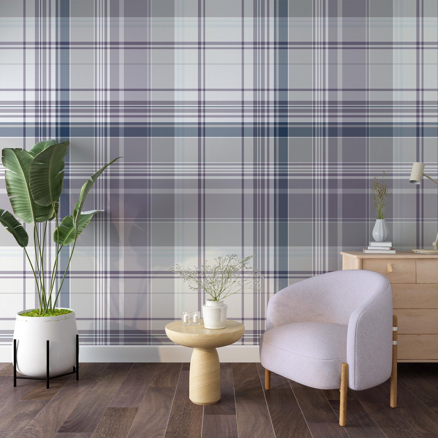 A stylish living room featuring a large plaid-patterned wallpaper in soft lilac, gray, and navy tones. The space includes a pale lavender accent chair, a round wooden side table with a small vase of flowers, a wooden cabinet with decor, and a tall green potted plant on dark wood flooring.