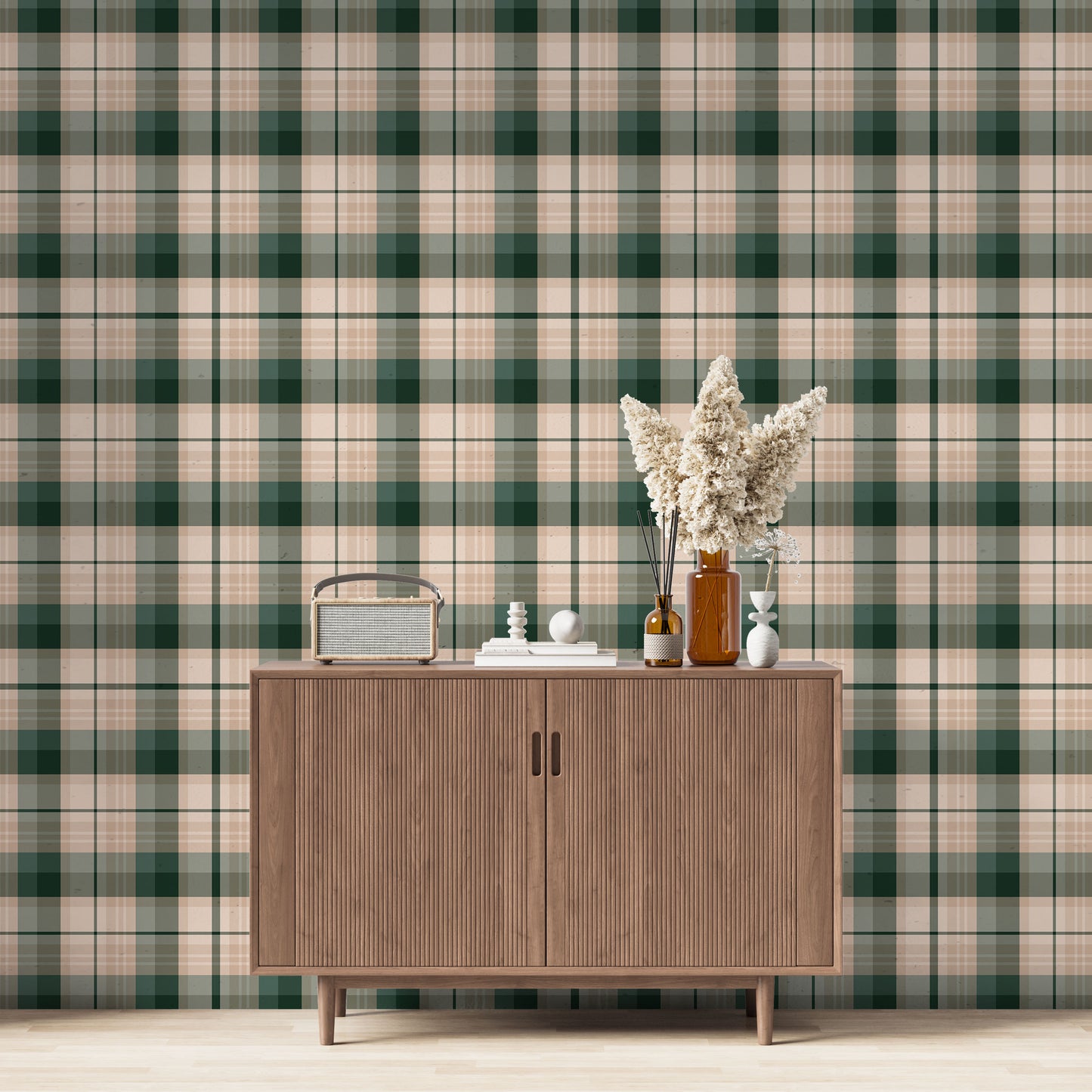 A modern living space featuring a deep forest green, sage green, and peach-tan plaid wallpaper accent wall. Centered against the wall is a mid-century modern wooden sideboard cabinet with vertical fluted doors and tapered legs. On top of the cabinet rests a vintage-style portable radio on the left, a small stack of white books with a geometric sphere on them in the center, and a collection of amber and white glass bottles to the right, filled with dried pampas grass and reed diffusers.