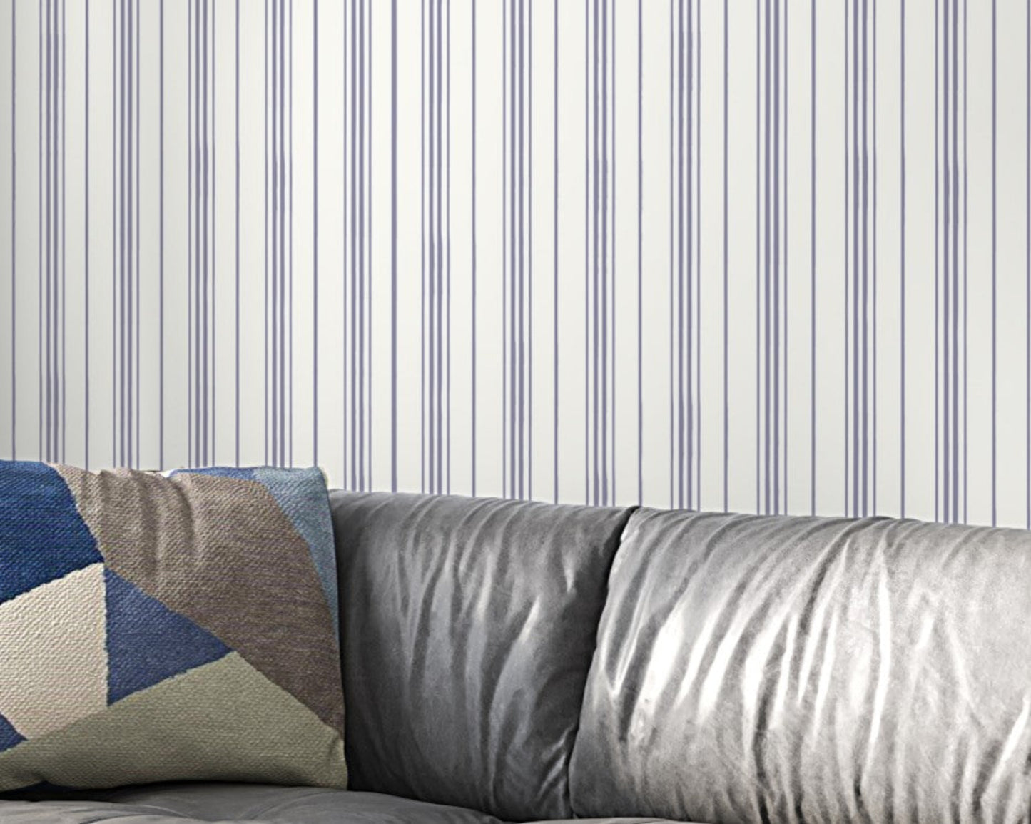 A gray leather sofa with a geometric-patterned throw pillow sits against a white wall decorated with thin vertical lavender-gray stripes. The minimalist interior features soft textures and a modern, neutral-toned design.
