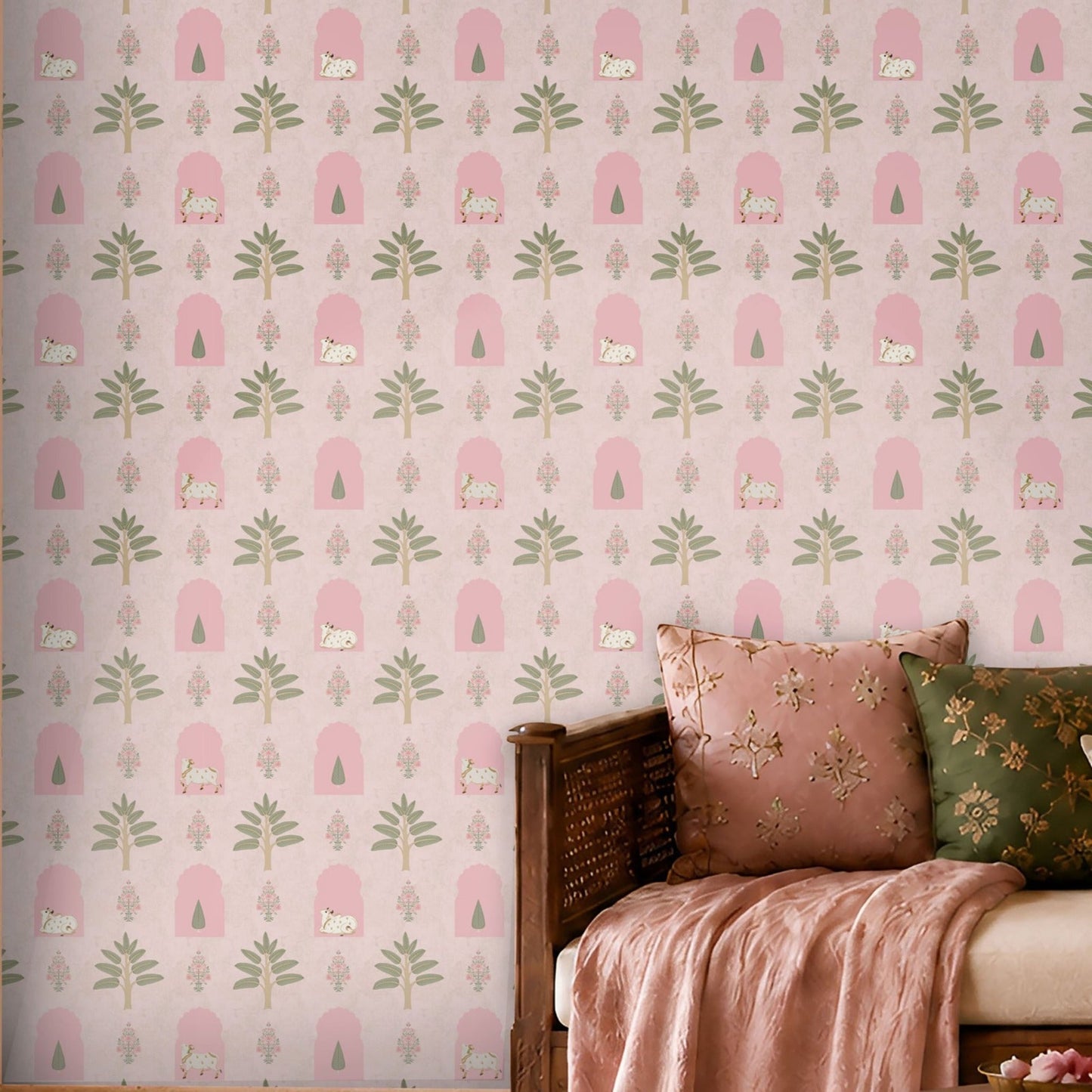 Blush pink Indian heritage wallpaper with floral, tree, arch and cow motifs, creating a charming accent wall in a cozy living room with wooden seating and pink cushions.