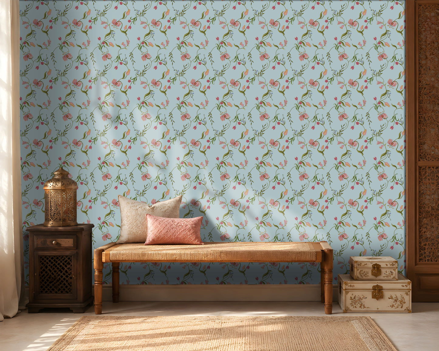 A cozy interior featuring a light blue floral wallpaper with delicate pink blossoms and green vines. A wooden woven bench with cream and blush pink cushions sits against the wall, flanked by a dark wooden side table holding a decorative metal lantern on the left and stacked cream-colored storage boxes on the right. Warm sunlight streams through sheer curtains, creating soft shadows across the wall and natural fiber rug, giving the room a calm, elegant atmosphere.