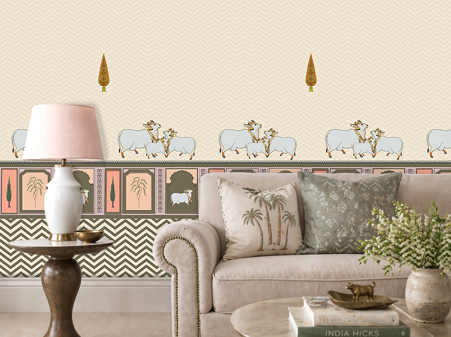 An elegant living room with off-white patterned wallpaper featuring subtle chevron textures, small tree motifs, and a decorative border with animal illustrations. In front is a beige upholstered sofa with cushions, including palm tree and geometric designs. A wooden side table holds a white lamp with a soft pink shade, while a round coffee table displays books, a small brass animal figurine, and a vase of flowers.