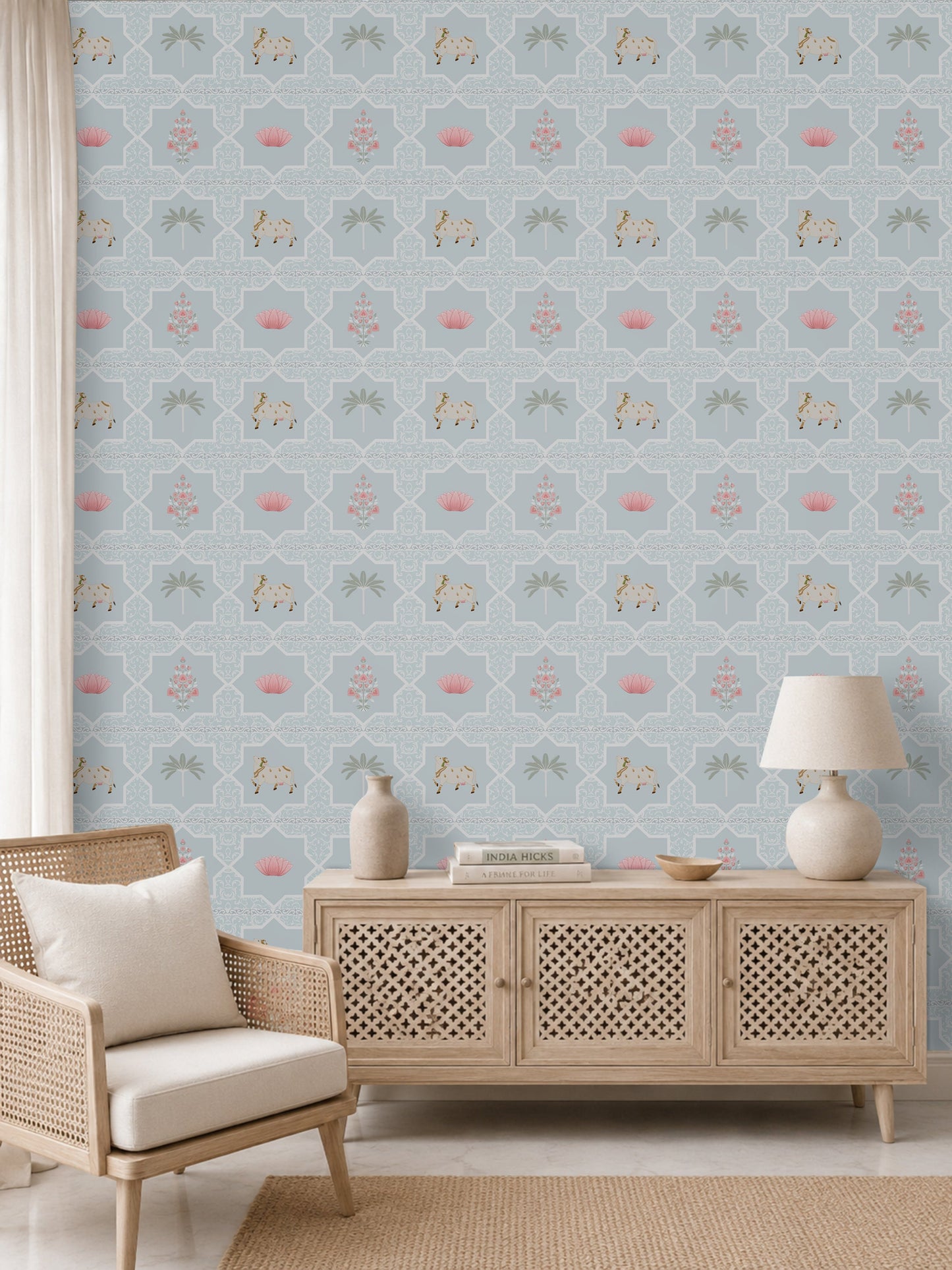 A modern bohemian living room or entryway featuring a powder blue Vraj Kamal Pichwai wallpaper patterned with cows and pink lotuses, styled with a light wood cane chair, a minimalist wooden console cabinet, and neutral decor.