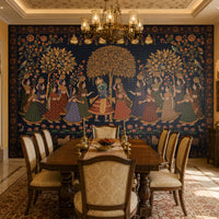 A spacious dining room showcases a dramatic wall-sized mural inspired by traditional Indian miniature paintings. At the center, Radha and Krishna stand beneath a golden flowering tree, surrounded by women in vibrant lehengas carrying decorative pots, with birds, floral vines, lotus flowers, and ornate borders enriching the composition. A long wooden dining table with upholstered chairs sits in the foreground, illuminated by an elegant chandelier. Warm beige walls, carved archways, polished ma