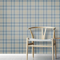 A cozy interior space features a classic plaid wallpaper in denim blue and sand tones.
The textured design displays a detailed grid pattern with intersecting blue, beige, and cream lines.
In front, a minimalist light-colored wooden wishbone chair with a woven seat is centered.
The simple setting is subtly completed by a small round side table holding a dark grey vase.
