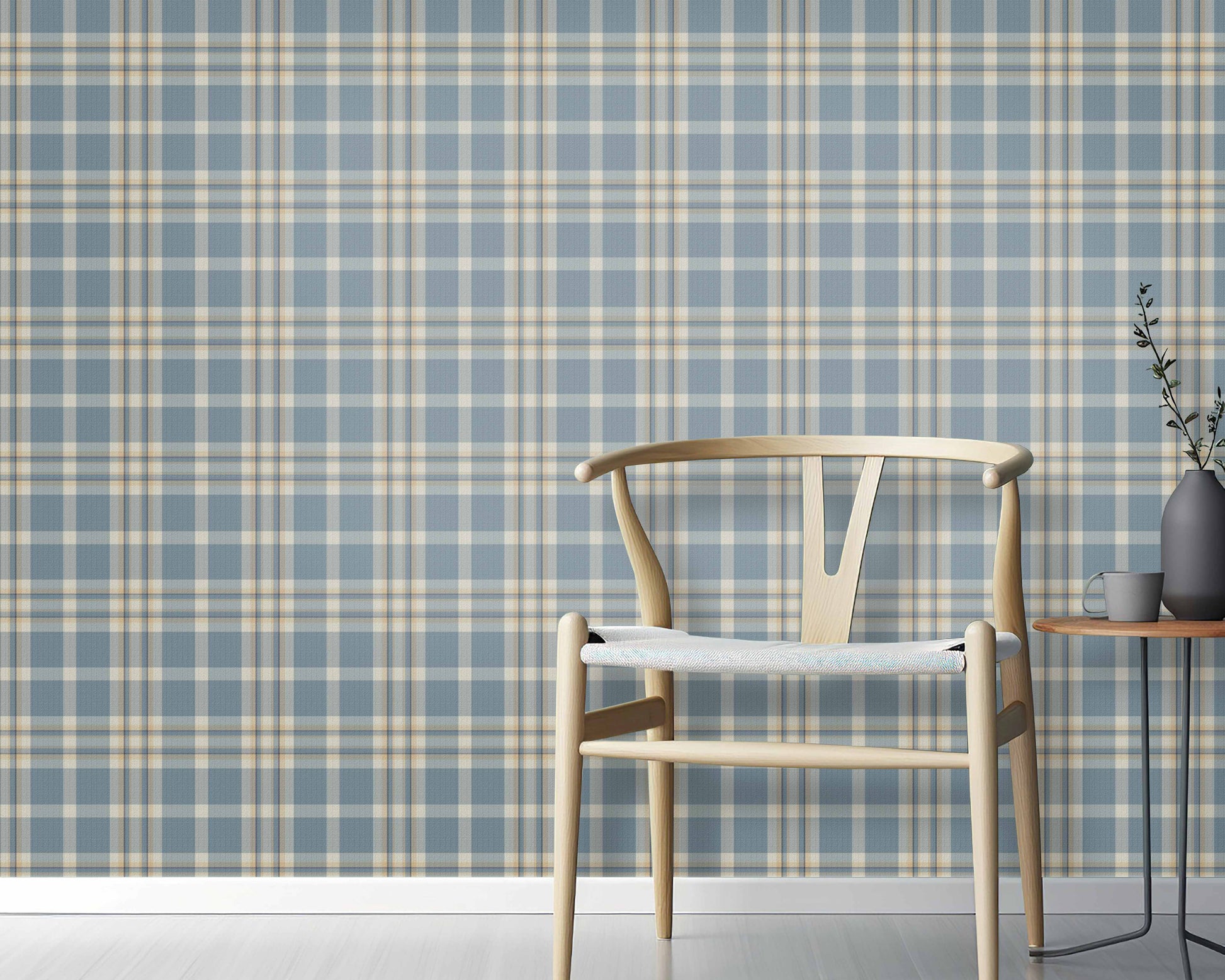 A cozy interior space features a classic plaid wallpaper in denim blue and sand tones.
The textured design displays a detailed grid pattern with intersecting blue, beige, and cream lines.
In front, a minimalist light-colored wooden wishbone chair with a woven seat is centered.
The simple setting is subtly completed by a small round side table holding a dark grey vase.