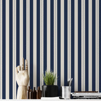 A structured workspace features a striking midnight blue and cream striped wallpaper.
The detailed design shows bold vertical navy bands and cream stripes with thin dotted lines.
In front, a clean white desktop is arranged with stationery, books, and a small green plant.
The contemporary, organized setup is uniquely accented by a decorative wooden hand model.