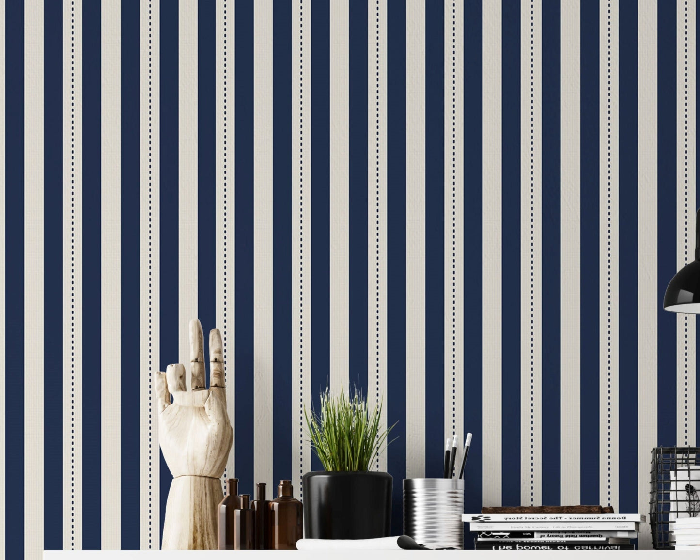 A structured workspace features a striking midnight blue and cream striped wallpaper.
The detailed design shows bold vertical navy bands and cream stripes with thin dotted lines.
In front, a clean white desktop is arranged with stationery, books, and a small green plant.
The contemporary, organized setup is uniquely accented by a decorative wooden hand model.