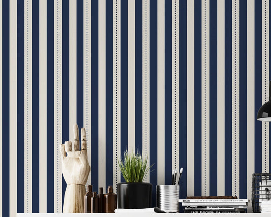 A structured workspace features a striking midnight blue and cream striped wallpaper.
The detailed design shows bold vertical navy bands and cream stripes with thin dotted lines.
In front, a clean white desktop is arranged with stationery, books, and a small green plant.
The contemporary, organized setup is uniquely accented by a decorative wooden hand model.