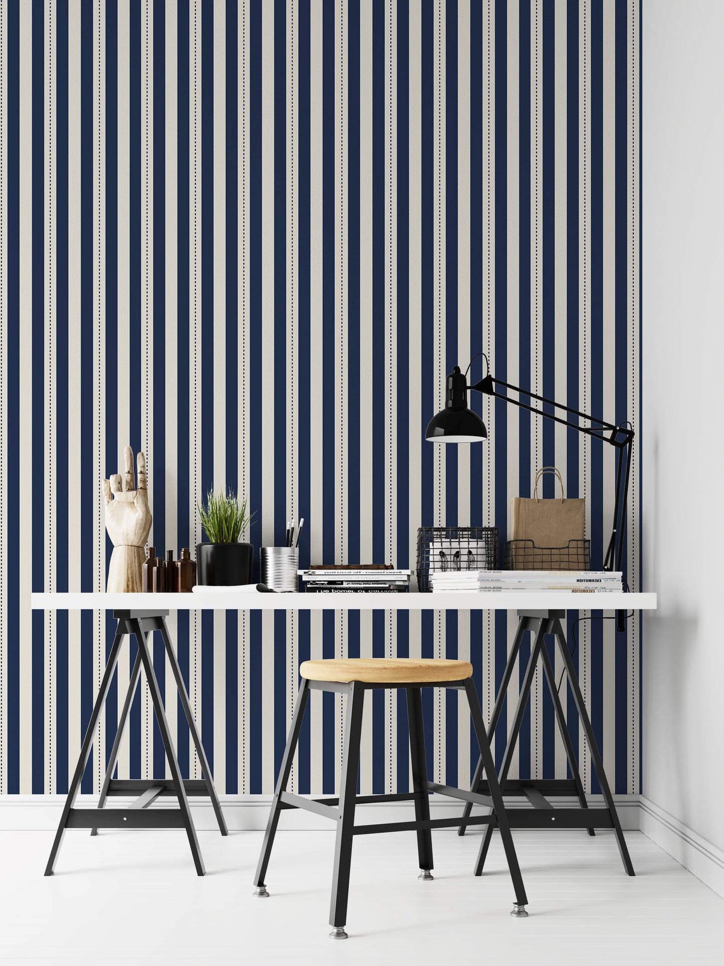 A structured workspace features a striking midnight blue and cream striped wallpaper.
The detailed design shows bold vertical navy bands and cream stripes with thin dotted lines.
In front, a clean white desktop is arranged with stationery, books, and a small green plant.
The contemporary, organized setup is uniquely accented by a decorative wooden hand model.