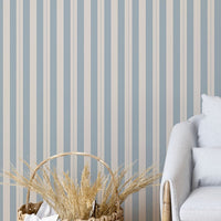 A modern living space features a soft slate blue and beige striped wallpaper.
The detailed design shows clean vertical blue bands alongside beige stripes with thin dotted lines.
In front, a comfortable light grey armchair with two matching pillows is centered nicely.
The relaxing setup is complete with a wooden tripod floor lamp and a woven storage basket.