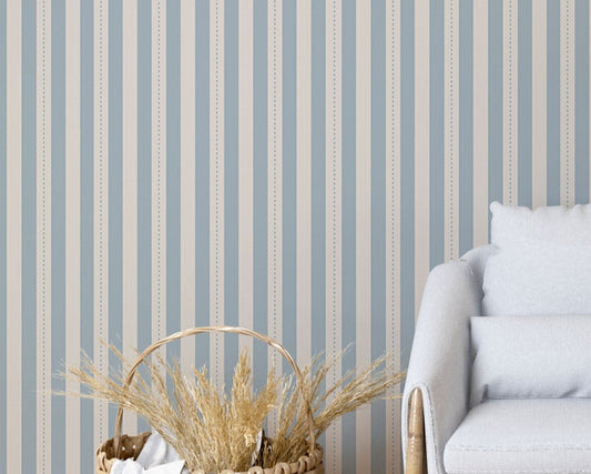 A modern living space features a soft slate blue and beige striped wallpaper.
The detailed design shows clean vertical blue bands alongside beige stripes with thin dotted lines.
In front, a comfortable light grey armchair with two matching pillows is centered nicely.
The relaxing setup is complete with a wooden tripod floor lamp and a woven storage basket.