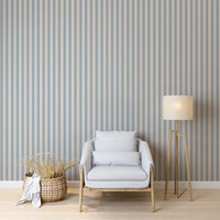 A modern living space features a soft slate blue and beige striped wallpaper.
The detailed design shows clean vertical blue bands alongside beige stripes with thin dotted lines.
In front, a comfortable light grey armchair with two matching pillows is centered nicely.
The relaxing setup is complete with a wooden tripod floor lamp and a woven storage basket.