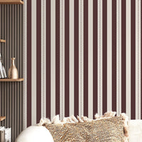 A creative workspace features a bold dark mahogany brown and beige striped wallpaper.
The detailed design shows clean vertical brown bands alongside beige stripes with thin dotted lines.
In front, a light wood desk table is neatly arranged with books, office supplies, and a clock.
The structured setup is grounded with a modern black stool and storage organizers.