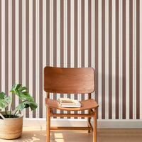 A warm, stylized workspace features a bold black and beige striped wallpaper.
The detailed design shows alternating vertical black bands and warm beige stripes.
In front, a light wood sawhorse desk table holds an assortment of books and office supplies.
The creative interior setup is completed with a black industrial pendant light and a stool.