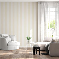 A sunlit living room features a striped wallpaper pattern.
The wallpaper colors are a pale cream beige and crisp white.
The design has wide, evenly spaced vertical stripes across the wall.
A large, rounded white armchair sits on the left side.
A light grey sectional sofa with pillows rests on the right.
The room is complete with natural wood flooring and white curtains.
