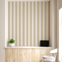 A compact workspace nook features a vertical striped wallpaper pattern.
The wallpaper colors are a muted almond beige and soft cream white.
The design has clean, evenly spaced vertical stripes filling the recessed wall.
A long, built-in light wood desk cabinet sits below the wallpaper.
The desk surface holds a small potted green plant and an open laptop.
A simple white wooden chair with a grey cushion is placed nearby.