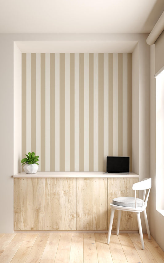 A compact workspace nook features a vertical striped wallpaper pattern.
The wallpaper colors are a muted almond beige and soft cream white.
The design has clean, evenly spaced vertical stripes filling the recessed wall.
A long, built-in light wood desk cabinet sits below the wallpaper.
The desk surface holds a small potted green plant and an open laptop.
A simple white wooden chair with a grey cushion is placed nearby.