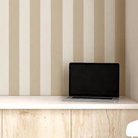 A compact workspace nook features a vertical striped wallpaper pattern.
The wallpaper colors are a muted almond beige and soft cream white.
The design has clean, evenly spaced vertical stripes filling the recessed wall.
A long, built-in light wood desk cabinet sits below the wallpaper.
The desk surface holds a small potted green plant and an open laptop.
A simple white wooden chair with a grey cushion is placed nearby.
