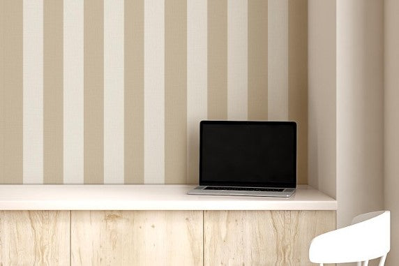 A compact workspace nook features a vertical striped wallpaper pattern.
The wallpaper colors are a muted almond beige and soft cream white.
The design has clean, evenly spaced vertical stripes filling the recessed wall.
A long, built-in light wood desk cabinet sits below the wallpaper.
The desk surface holds a small potted green plant and an open laptop.
A simple white wooden chair with a grey cushion is placed nearby.