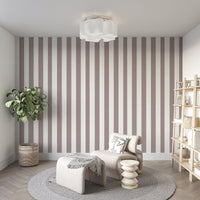 A modern living room features a muted taupe and off-white striped wallpaper.
The design showcases clean, evenly spaced vertical lines covering the main back wall.
In front, a stylish cream-colored lounge chair and matching ottoman sit on a round rug.
The minimalist space is complete with a large potted plant and a light wood bookshelf.