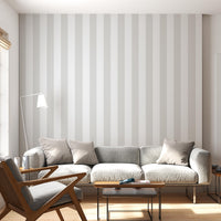 A bright, minimalist living room features a soft cloud whisper grey and white striped wallpaper.
The elegant design shows wide vertical grey and white bands repeating evenly across the main wall.
In front, a large grey sofa with neutral toss pillows sits next to a wooden mid-century armchair.
The calming space is nicely complemented by small wooden side tables and a slender white floor lamp.