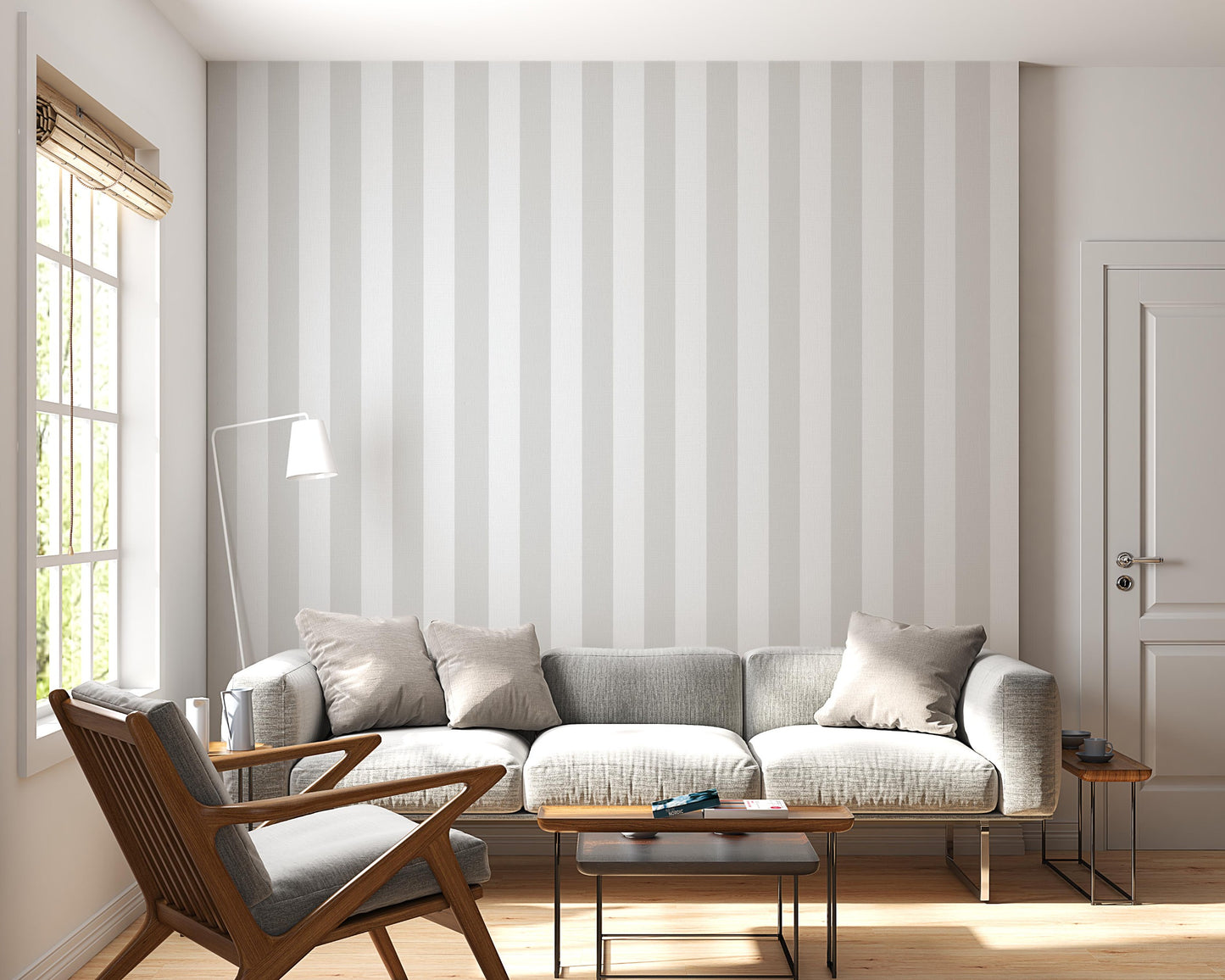 A bright, minimalist living room features a soft cloud whisper grey and white striped wallpaper.
The elegant design shows wide vertical grey and white bands repeating evenly across the main wall.
In front, a large grey sofa with neutral toss pillows sits next to a wooden mid-century armchair.
The calming space is nicely complemented by small wooden side tables and a slender white floor lamp.