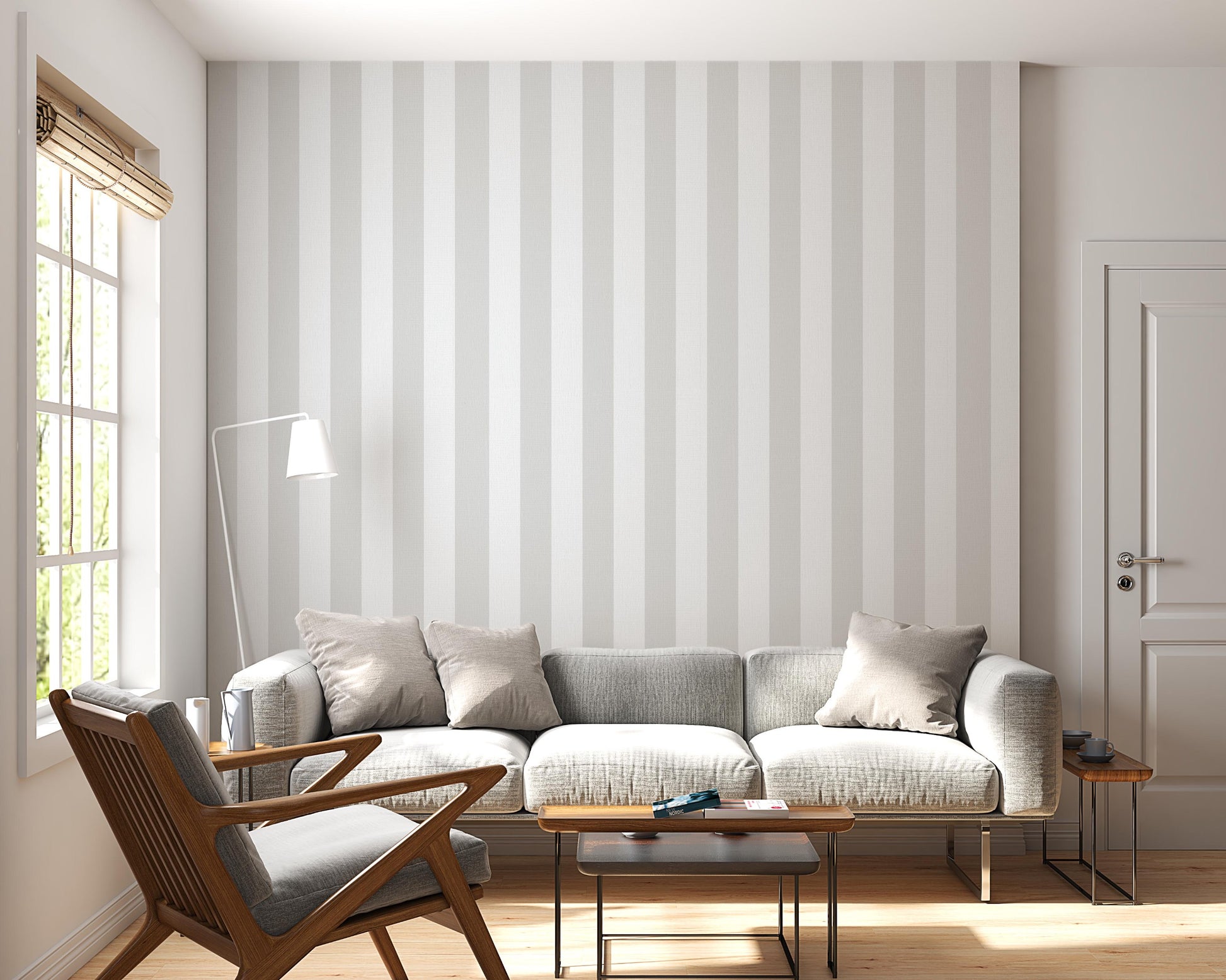 A bright, minimalist living room features a soft cloud whisper grey and white striped wallpaper.
The elegant design shows wide vertical grey and white bands repeating evenly across the main wall.
In front, a large grey sofa with neutral toss pillows sits next to a wooden mid-century armchair.
The calming space is nicely complemented by small wooden side tables and a slender white floor lamp.