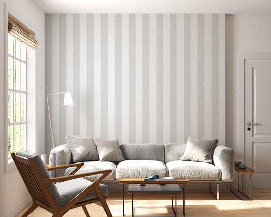 A bright, minimalist living room features a soft cloud whisper grey and white striped wallpaper.
The elegant design shows wide vertical grey and white bands repeating evenly across the main wall.
In front, a large grey sofa with neutral toss pillows sits next to a wooden mid-century armchair.
The calming space is nicely complemented by small wooden side tables and a slender white floor lamp.