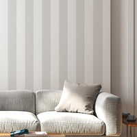 A modern living room features a muted taupe and off-white striped wallpaper.
The design showcases clean, evenly spaced vertical lines covering the main back wall.
In front, a stylish cream-colored lounge chair and matching ottoman sit on a round rug.
The minimalist space is complete with a large potted plant and a light wood bookshelf.