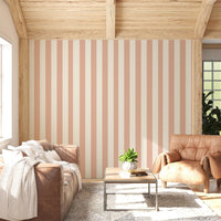 A sunlit living room features a warm honey-taupe and cream striped wallpaper.
The design showcases clean, evenly spaced vertical stripes across the central wall.
In front, a brown leather sofa and matching armchair frame a small coffee table.
The rustic space is complete with a textured area rug and a light wood beamed ceiling.