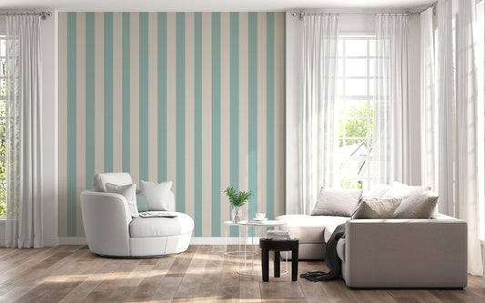 A sunlit living room features a soft sage blue and warm cream striped wallpaper.
The design showcases clean, evenly spaced vertical stripes filling the central accent wall.
In front, a rounded white armchair and a light grey sectional sofa face each other.
The bright, modern space is complete with natural wood flooring and sheer white curtains.