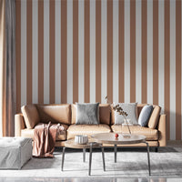 A modern living room features a warm cocoa brown and off-white striped wallpaper.
The design showcases clean, evenly spaced vertical stripes filling the central wall.
In front, a tan leather sofa with decorative pillows frames a round coffee table setup.
The stylish space is complete with a large area rug and an accent bar stool on the right.