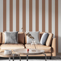 A modern living room features a warm cocoa brown and off-white striped wallpaper.
The design showcases clean, evenly spaced vertical stripes filling the central wall.
In front, a tan leather sofa with decorative pillows frames a round coffee table setup.
The stylish space is complete with a large area rug and an accent bar stool on the right.