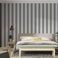A modern bedroom features a simple grey and white striped wallpaper.
The design showcases clean, evenly spaced vertical stripes filling the main wall.
In front, a fully dressed bed with a fabric headboard centers the cozy setup.
The styled space is finished with a wooden bench, a side table, and a green plant.