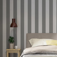 A modern bedroom features a simple grey and white striped wallpaper.
The design showcases clean, evenly spaced vertical stripes filling the main wall.
In front, a fully dressed bed with a fabric headboard centers the cozy setup.
The styled space is finished with a wooden bench, a side table, and a green plant.