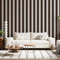 A stylish living room features a dark chocolate brown and crisp white striped wallpaper.
The design showcases clean, evenly spaced vertical stripes across the expansive back wall.
In front, a plush cream-colored sofa and matching armchair frame sleek round coffee tables.
The modern, high-contrast space is complete with a geometric area rug and potted green plants.