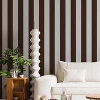A stylish living room features a dark chocolate brown and crisp white striped wallpaper.
The design showcases clean, evenly spaced vertical stripes across the expansive back wall.
In front, a plush cream-colored sofa and matching armchair frame sleek round coffee tables.
The modern, high-contrast space is complete with a geometric area rug and potted green plants.