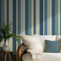 A coastal themed living area features a bold stormy tides blue and cream striped wallpaper.
The detailed design shows alternating thick and thin vertical stripes in varying shades of blue and tan.
In front, a woven rattan sofa with white cushions and blue accent pillows is centered nicely.
The refreshing, tropical setup is flanked by lush green potted plants in woven baskets on both sides.