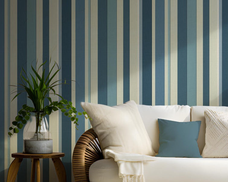 A coastal themed living area features a bold stormy tides blue and cream striped wallpaper.
The detailed design shows alternating thick and thin vertical stripes in varying shades of blue and tan.
In front, a woven rattan sofa with white cushions and blue accent pillows is centered nicely.
The refreshing, tropical setup is flanked by lush green potted plants in woven baskets on both sides.