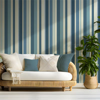 A coastal themed living area features a bold stormy tides blue and cream striped wallpaper.
The detailed design shows alternating thick and thin vertical stripes in varying shades of blue and tan.
In front, a woven rattan sofa with white cushions and blue accent pillows is centered nicely.
The refreshing, tropical setup is flanked by lush green potted plants in woven baskets on both sides.