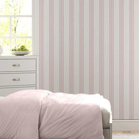 A bright bedroom features a soft pink and white vertically striped wallpaper.
The detailed design shows thin, evenly spaced pink stripes running down a white base.
In front, a neatly made bed with a light pink comforter rests near a matching nightstand.
The relaxing space is complete with two light grey dressers, large windows, and a rug.