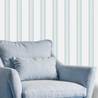A bright sitting area features a clean coastal dune blue and white striped wallpaper.
The detailed design shows thin blue and beige vertical stripes repeating down a light base.
In front, a plush light blue armchair with a matching lumbar pillow rests comfortably.
The relaxing space is complete with a small wooden side table holding a mug and plant.