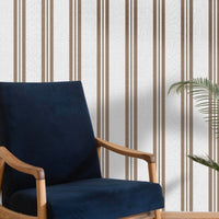 A bright seating area features a warm rustic mocha brown and white striped wallpaper.
The detailed design shows clean brown vertical lines repeating across a textured light base.
In front, a classic dark blue velvet armchair with a wooden frame sits comfortably.
The simple, inviting space is naturally accented with a leafy green potted plant nearby.