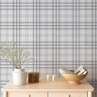 A serene indoor space features a soft stormy lavender and white plaid wallpaper.
The detailed design shows thin purple-grey lines intersecting to form clean grids across a light base.
In front, a light wooden tabletop holds books, a wooden bowl, and small candles.
The calming setup is beautifully accented with a white vase containing delicate white flowers.