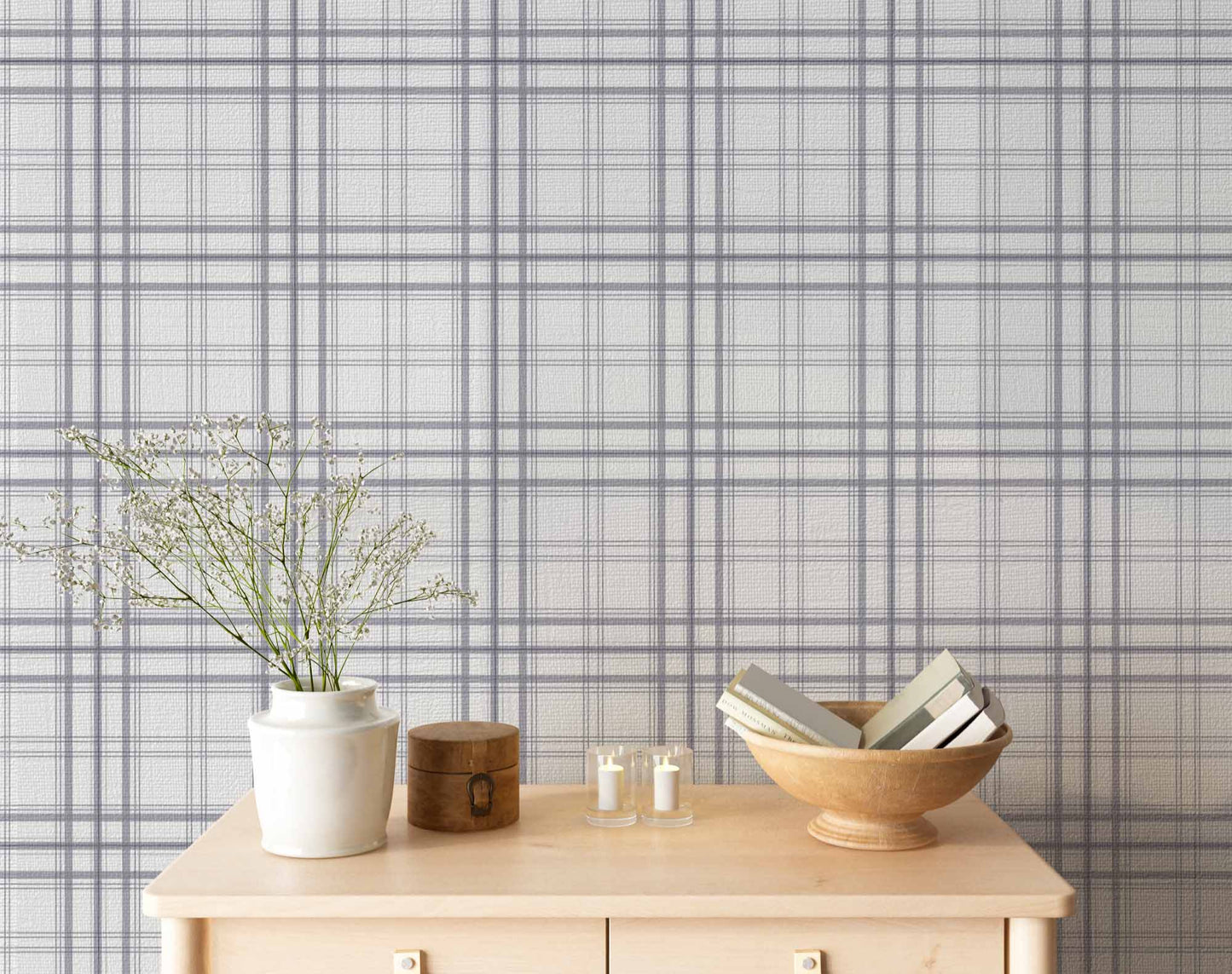 A serene indoor space features a soft stormy lavender and white plaid wallpaper.
The detailed design shows thin purple-grey lines intersecting to form clean grids across a light base.
In front, a light wooden tabletop holds books, a wooden bowl, and small candles.
The calming setup is beautifully accented with a white vase containing delicate white flowers.