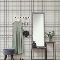 A warm interior setup features a soft faded olive green and grey plaid wallpaper.
The detailed design shows thin dark lines intersecting to form clean grids across a light olive base.
In front, a light wood media console with woven rattan doors is centered nicely.
The relaxing space is accented with a wooden armchair, decorative pottery, and dried botanical stalks.