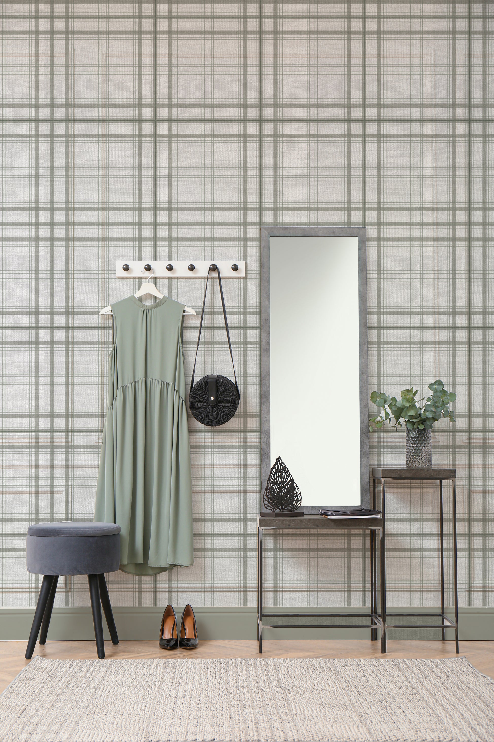 A warm interior setup features a soft faded olive green and grey plaid wallpaper.
The detailed design shows thin dark lines intersecting to form clean grids across a light olive base.
In front, a light wood media console with woven rattan doors is centered nicely.
The relaxing space is accented with a wooden armchair, decorative pottery, and dried botanical stalks.
