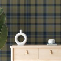 A cozy room features a dark olive green and navy blue plaid wallpaper.
The detailed design shows intersecting dark stripes over a muted green base.
In front, a light wood chest of drawers stands next to a small matching side table.
The rustic space is accented with large potted plants and minimalist wall shelves.