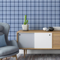 A contemporary living room with blue plaid wallpaper creating a textured backdrop. The space features a gray upholstered armchair, a mid-century modern wooden sideboard with decorative objects and books, and a large potted indoor tree on dark wood flooring.